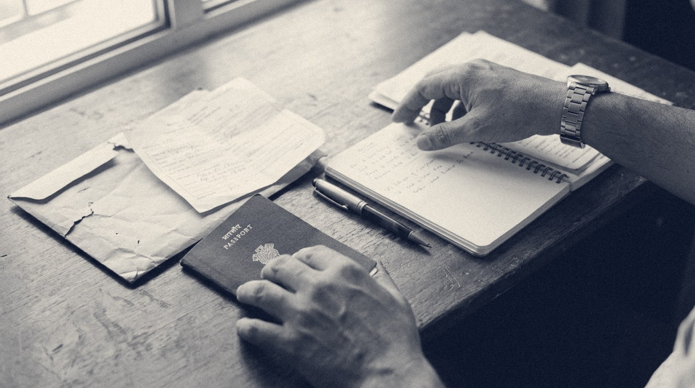 Hands sorting paperwork on a wooden desk: a dark Indian passport, a folded form, a worn manila envelope and a notebook with handwritten notes.